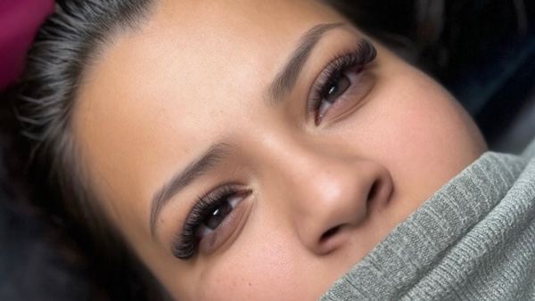 Close-up of a woman's eyes with voluminous eyelash extensions, peering over a grey fabric.